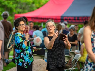 Two alumni looking at a phone, with a red and black awning and several people in the background