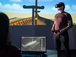 Person in VR gear with image of the VR scene of a longboat in the background and person on computuer with data and black and white image of the boat
