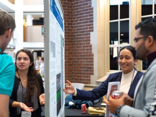Two women present their research posters in a busy atrium. They stand on either side of a bulletin board, and are gesticulating toward their posters while speaking to listeners in the foreground.