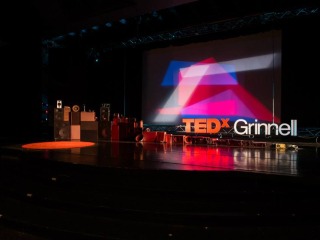 Dark stage with podium and background lit in reds. Set pieces reads TEDx Grinnell