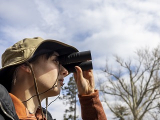 A woman in a beige, wide-brim hat holds a pair of binoculars to her eyes and gazes off camera. Behind her, leafless trees are backed by a blue, cloudy sky.