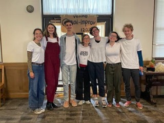 Seven people in casual clothes stand in front of a double-door displaying a cardboard sign that includes the words venmo and improve