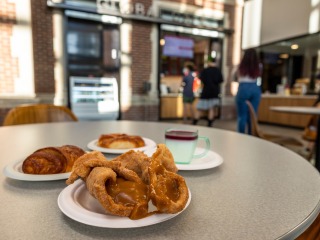 A tasty array of pastries and a hot drink sit on a table while a line of people queue for the cafe in the background