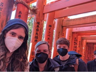A selfie taken by three people in winter jackets and face masks. behind them are red, wooden gates inscribed with Japanese calligraphy.