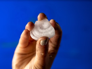 A frozen hailstone is held against a blue background by a white hand. Visible on the hail's surface are rings of clear and cloudy ice.