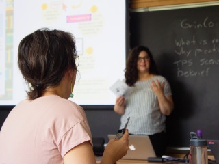 A faculty members leads a presentation next to a PowerPoint screen; a faculty member looks on.