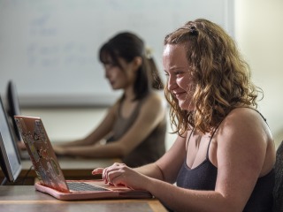 Kailee, a young woman with curly brown hair, smiles while working at her laptop computer.