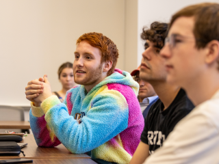 A ginger haired student with a colorful pink, blue, green sweater, hands clasped, looks off screen with a slight smile.