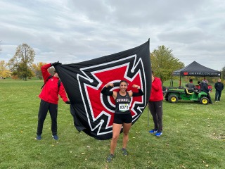 A young woman runner poses in front of Grinnell's Honor G flag