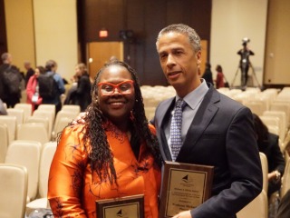 A Black women with red glasses and a bright orange shirt smiles alongside a gray-haired man wearing a suit.