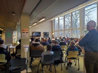 Rows of seats are occupied by people in the first floor lounge of the library. Someone stands on the right side of the room. 