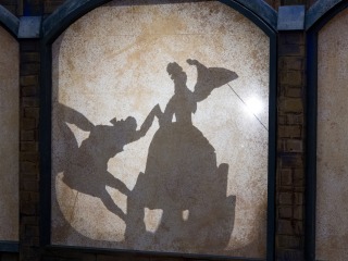 A grainy, light beige bacground split into three sides. In the middle square, a shadow of a person in a huge dress being held by a shadow of a hand. On their left, a shadow of someone bowing in formal duke-like clothes.