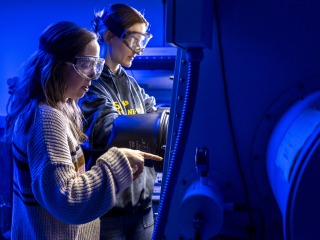 In a dark laboratory, a professor with long blond hair points inside a protected hood, while a student reaches inside the hood to manipulate an experiment.