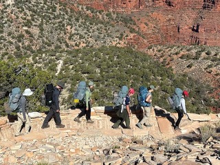 6 hikers with camping gear travel a stone-lined path. Scrub land to their left leads to red stone cliffs 