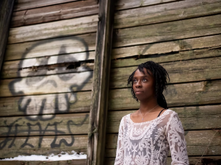 A young woman poses against a rough wooden wall painted with graffiti 