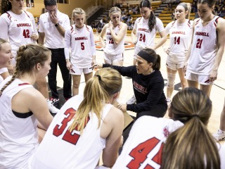 The women's basketball team dressed in white uniforms surround their coach, in black, before a game