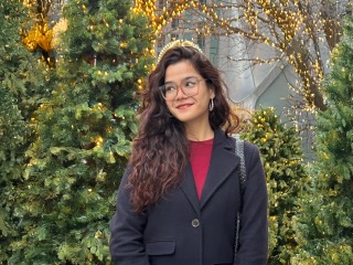 Meghna stands in front of a number of trees, some lit with white holiday lights
