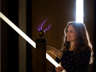 Charlotte Christensen holding a long cylindrical light with a metal sphere emitting purple cracks of light sits in front