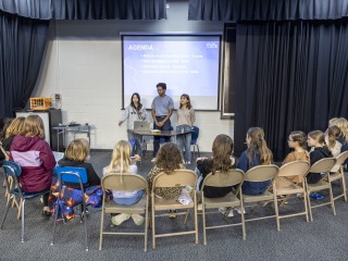 Three college students stand facing a semicircle of 15 middle school girls sitting in folding chairs