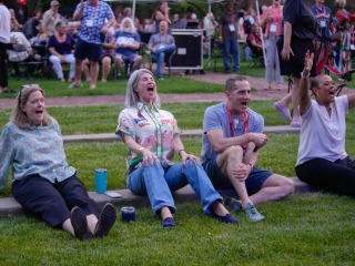 Four alums sitting on the grass cheering for the on stage performance.