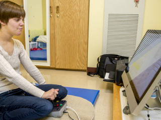 Jennifer Brooks ’15 sits on a blue gymnastics mat, facing a Mac on a table about 4 inches off the floor.