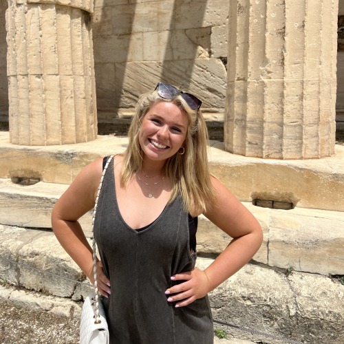 A young woman in sunglasses poses in front of ancient ruins in Greece.