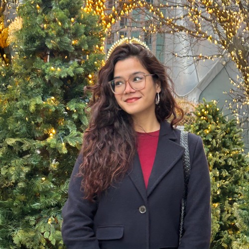 Meghna stands in front of a number of trees, some lit with white holiday lights