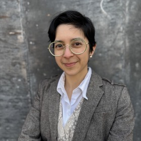 Professor Núñez in a tweed suit, standing in front of a gray metal wall