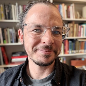 Portrait of a white transgender professor in front of his bookshelf