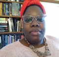 A Black woman with glasses wears a red headwrap, pink sweater and pink chunky necklace looks straight at the camera. She stands in front of a bookshelf.