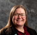 White, cis-gender woman with long hair and glasses, burgundy shirt, black jacket, and owl pin