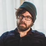 Farrar has a relaxed expression and sports a beard, glasses, and dark green beanie.