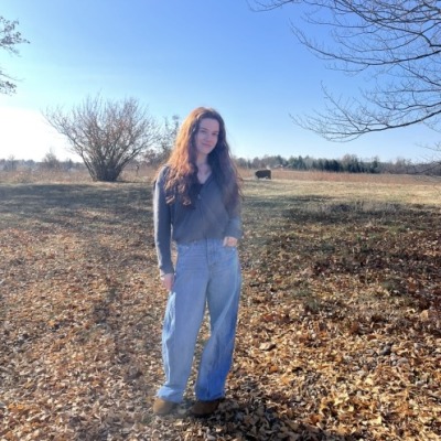 A woman stands in a leaf-covered field, in front of a wide blue sky