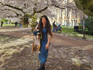 Cassey stands under a blooming cherry blossom tree and holds her dog. She is wearing a denim dress and has her long hair down.