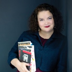 A woman with curly hair holds books. She smiles at the camera.