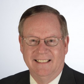 An older white man smiles at the camera. He wears glasses and a black suit.