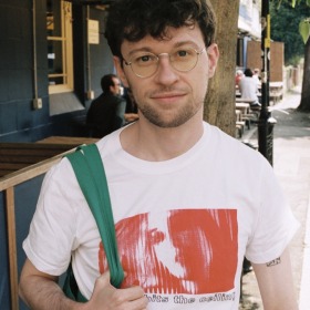 Man with short dark hair and round glasses wearing a white t-shirt with a red logo