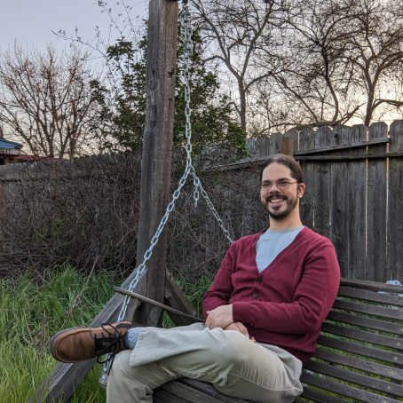 A man in a red cardigan and khakis sits on a wooden bench in his back yard. His legs are crossed and he is smiling.