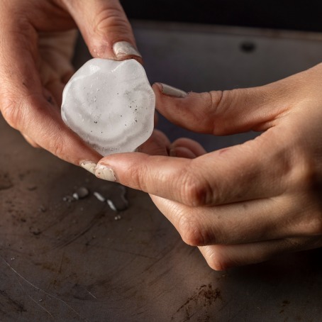 Two hands hold a half of a hailstone. Cloudy layers are visible on the halve's flat surface.