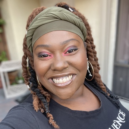 A Black women with lightbrown locs smiles at the camera. She is wearing bright pink eyeshadow and a black shirt.
