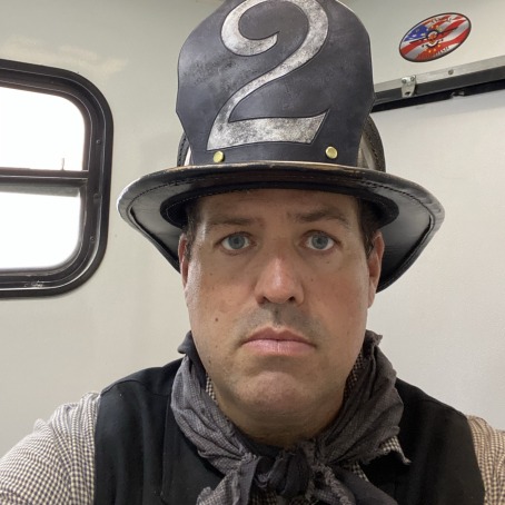 A man, wearing a brown tall hat with the number 2 printed, stares into the camera for a selfie. He also wears a scarf and vest.