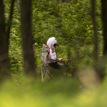 A student in a white jacket sitting in a forest as seen through trees.