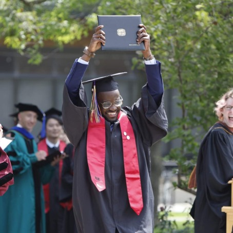 MJ Ketcha wearing black graduation robes with a red stole and holding a diploma above his head.