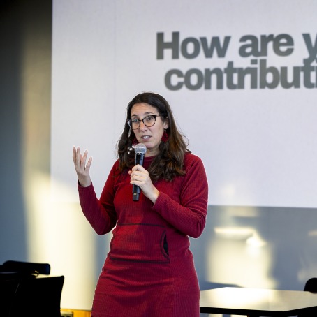 Daniela Papi-Thornton wearing a red sweater and holding a microphone in front of a projected slide that reads "How are you a contribution?"
