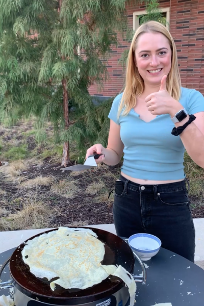 Catherine standing outside holding a metal turner and giving a thumbs up with a successfully flipped crepe in the electric griddle before her.