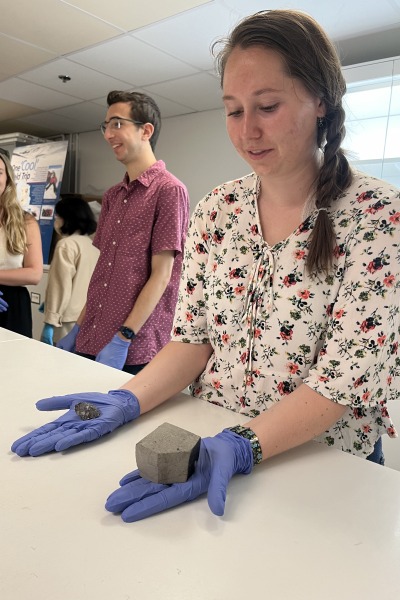 Emma Stefanacci  wears blue gloves with a small irregular meteorite in one hand and a larger, palm sized cube of rock in the other.