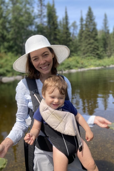A smiling woman carrying a smiling baby with a stream and forest behind her 
