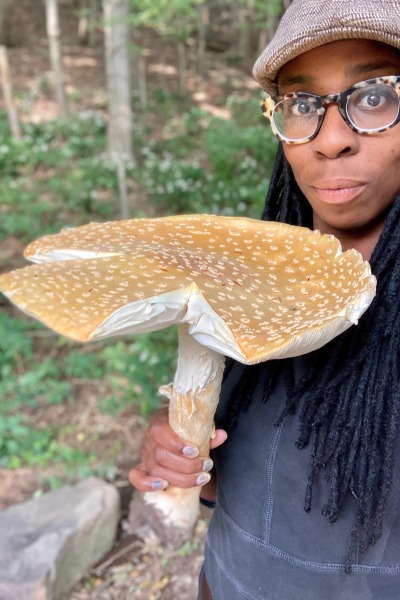 A woman in a hat and glasses holds an enormous mushroom