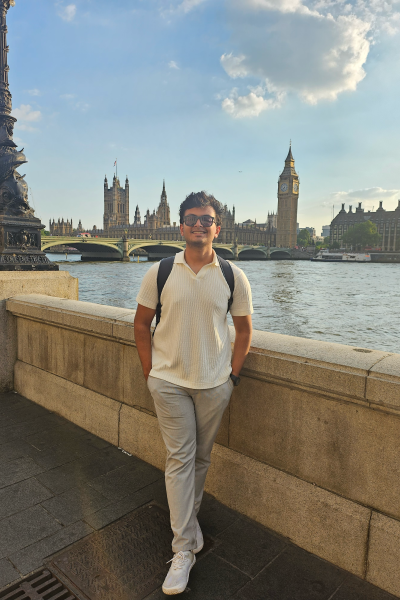 Parikshit stands on the banks of the Thames River in London with the Tower of London and Parliament behind him