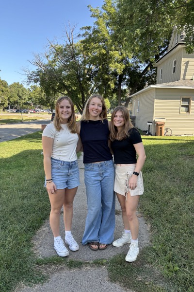 Three young women stand arm in arm on a sidewalk with green grass and trees all around